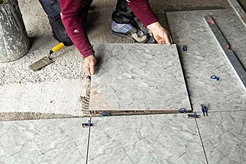 Worker installing a large grey floor tile on adhesive using spacers for alignment.