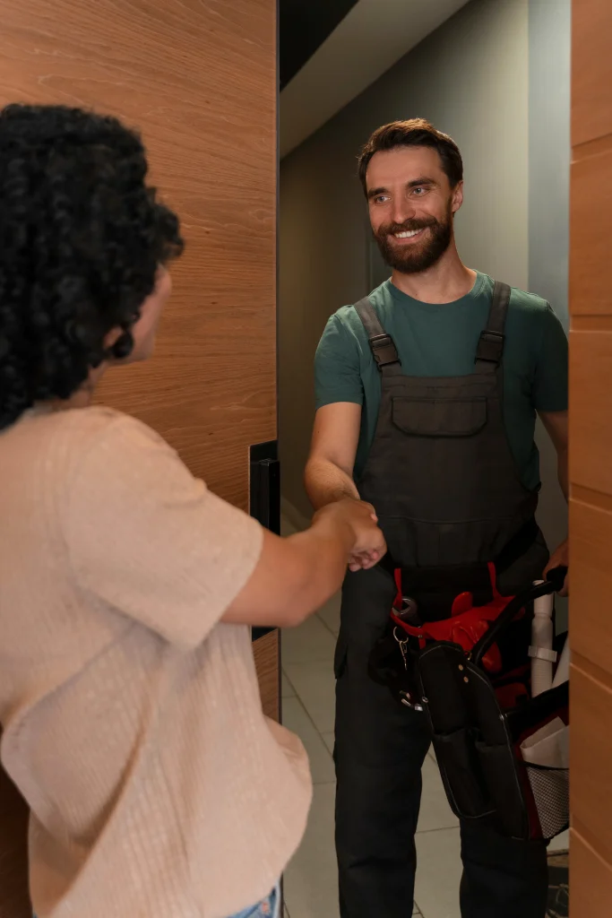A handyman standing at a client's front door, smiling and shaking hands while holding a tool bag.