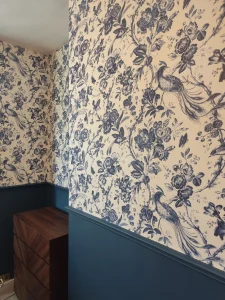 A room featuring blue and white floral bird wallpaper above a dark teal wainscoted wall with a walnut dresser.