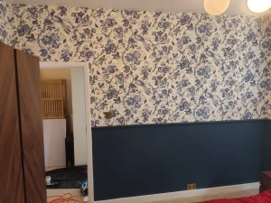 A room wall with bluebird-patterned wallpaper on the top half and dark teal paint on the bottom half, including a doorway and walnut furniture.